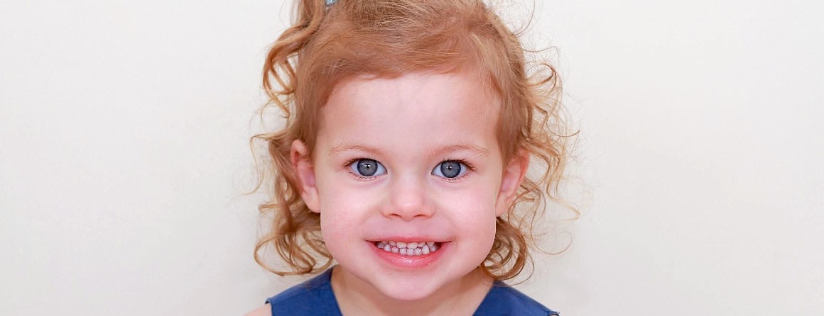 A little girl with pale skin, strawberry blonde hair and a blue dress smiles at the camera.